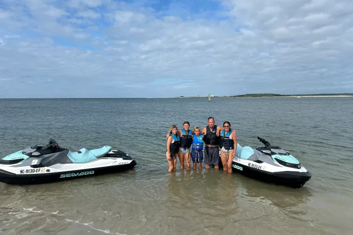 Group of people in life jackets standing between two jet skis on a beach.
