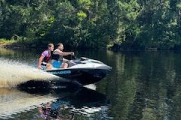 Two people riding a jet ski on a sunny day with trees in the background.