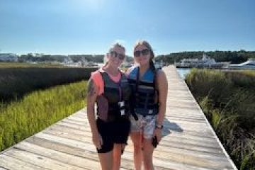 Two people in life jackets on a wooden dock under a clear blue sky.