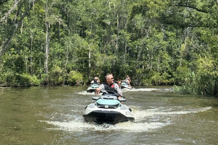 People ride jet skis on a river surrounded by lush green trees and clear skies.