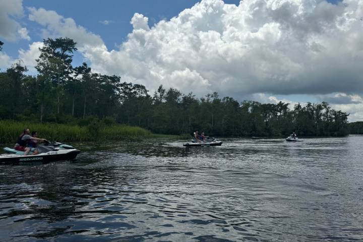Three people riding jet skis on a river with trees and clouds in the background.