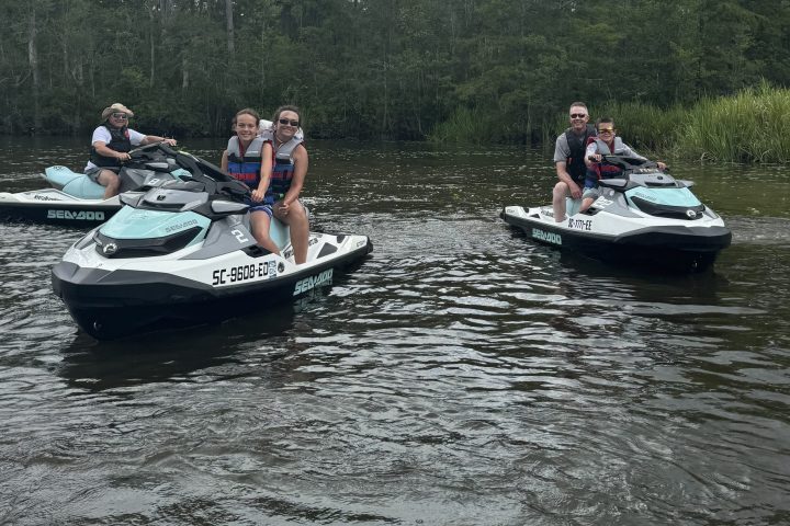 People on jet skis in a river surrounded by trees under a cloudy sky.