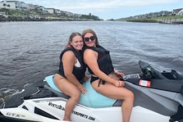 Two people on a jet ski wearing life jackets on a cloudy day near a shoreline.