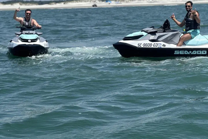 Two people on jet skis giving thumbs up on the ocean near a sandy beach under a cloudy sky.