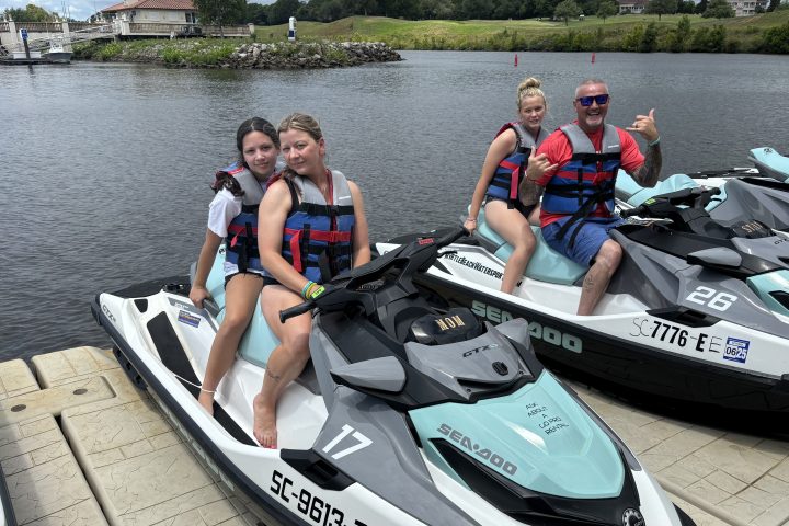 People wearing life jackets sit on jet skis at a dock with water and buildings in the background.