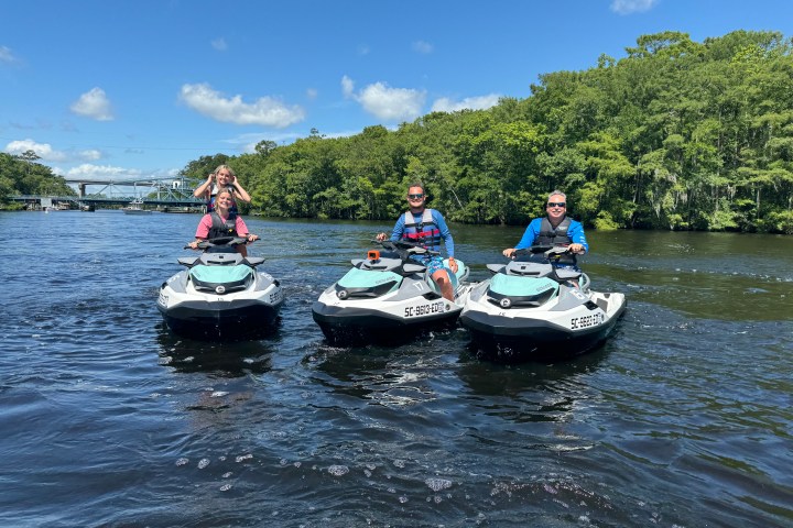 Three people on jet skis on a river with a bridge and trees in the background.