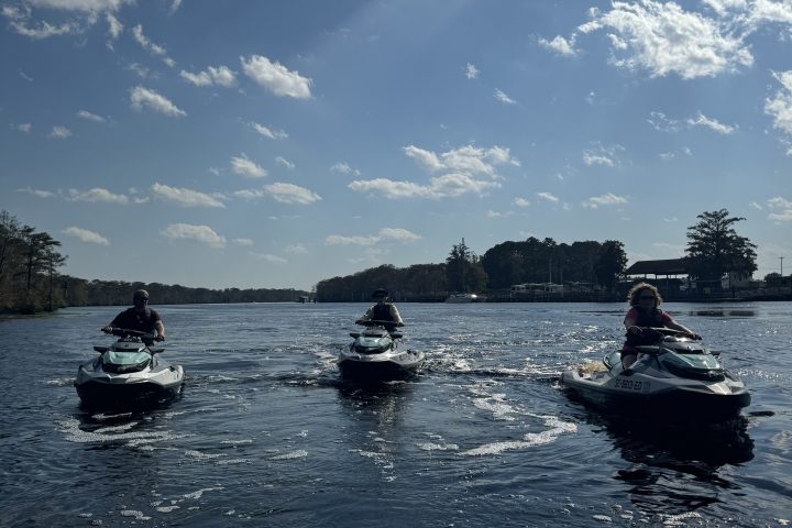 Three people on jet skis riding on a wide river under a sunny, partially cloudy sky.