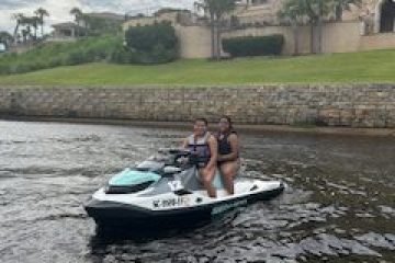 Two people on a jet ski in front of a large house by the water.