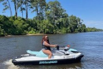 Person riding a jet ski on a river with trees and clear blue sky in the background.