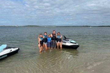 Group of five people in life vests standing in shallow water near jet skis.