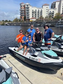 Group of six people posing on and around a jet ski by a marina, with buildings in the background.
