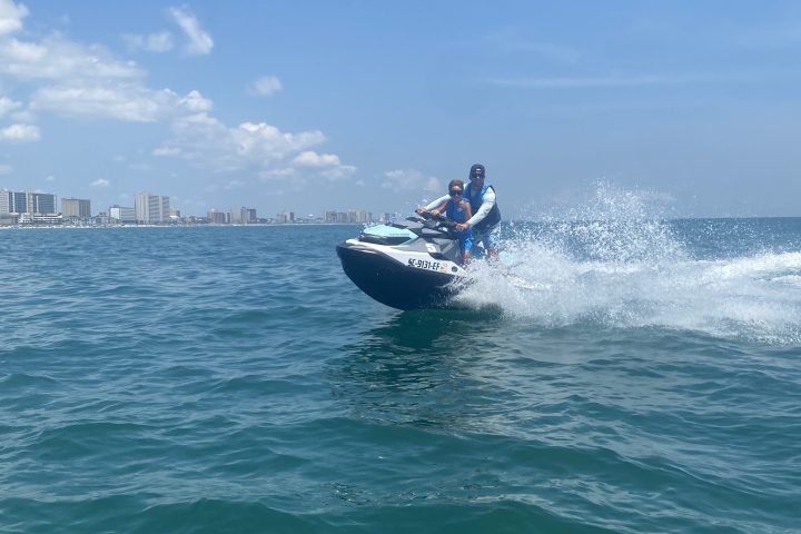 Two people riding a jet ski on the ocean with city skyline in the background.