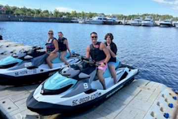 Two pairs of people on jet skis docked by a marina with boats in the background.