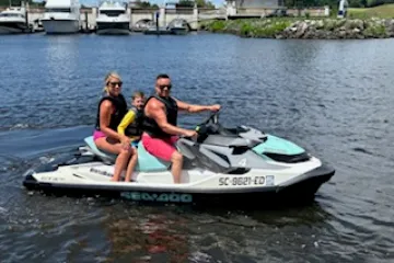 Three people on a jet ski in a marina with boats and buildings in the background.