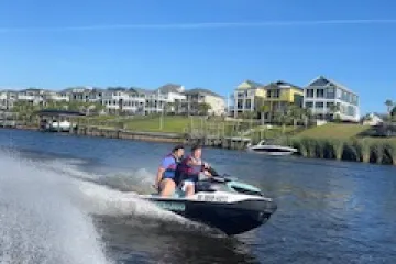 Two people on a jet ski riding on a waterway with houses in the background.