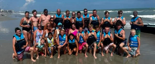 Group of people wearing life vests posing on a beach with waves in the background.