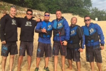Six men in matching blue and black gear pose on a sandy beach with trees in the background.