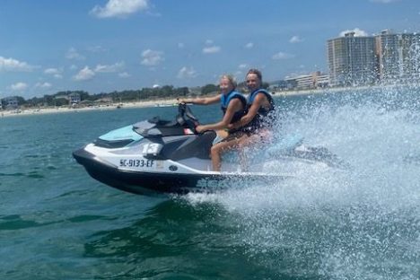 Two people ride a jet ski on the ocean with a cityscape in the background.