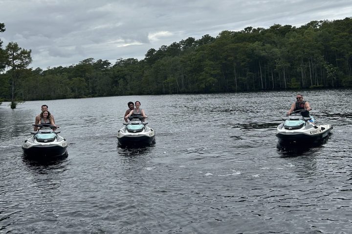 Three people on jet skis riding on a calm lake under a cloudy sky.