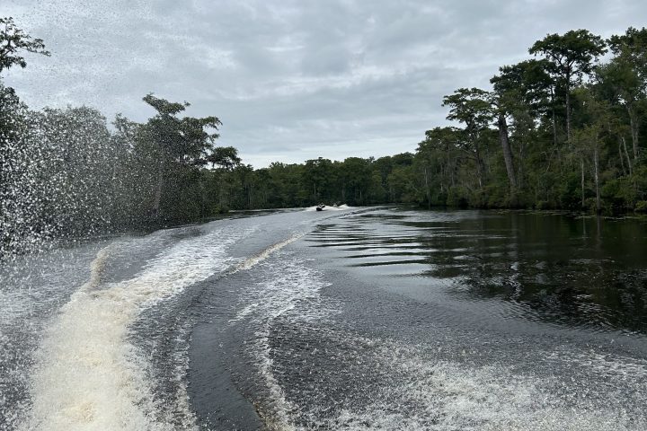 Boat wake on a river with trees on the sides and a cloudy sky.