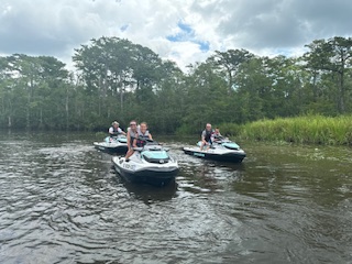 Three people riding jet skis on a river surrounded by lush trees under cloudy skies.
