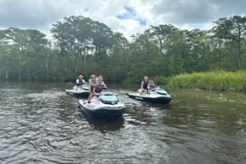 Group of people riding jet skis on a river with trees in the background under a cloudy sky.