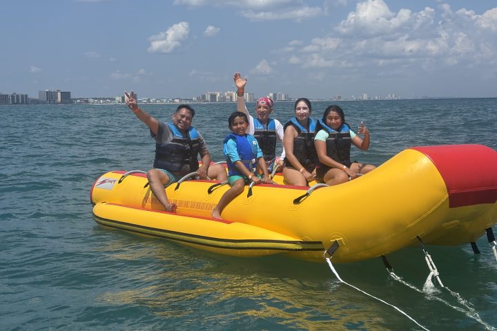 Five people on a yellow inflatable banana boat in the ocean, smiling and waving.