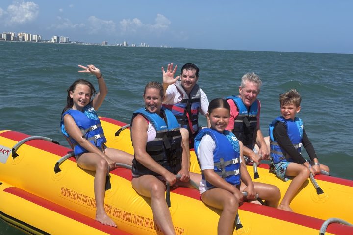 Group of six people on a yellow inflatable raft in the ocean, wearing blue life jackets on a sunny day.