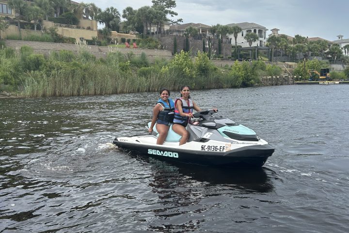 Two people on a jet ski in a body of water, houses and trees in the background.