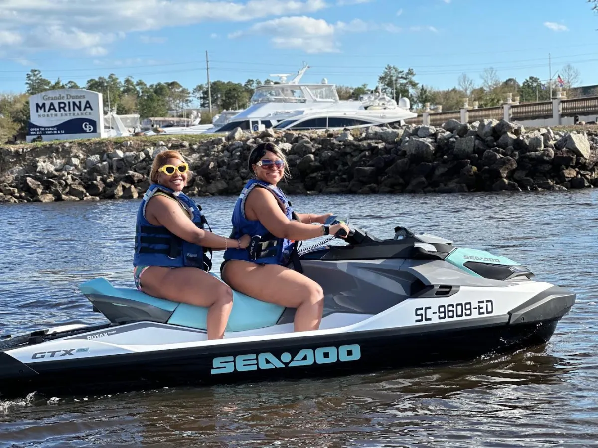 Two people on a jet ski in a marina with boats and a sign in the background.