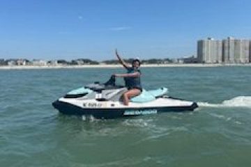 Person on a jet ski waving in coastal waters with buildings in the background.