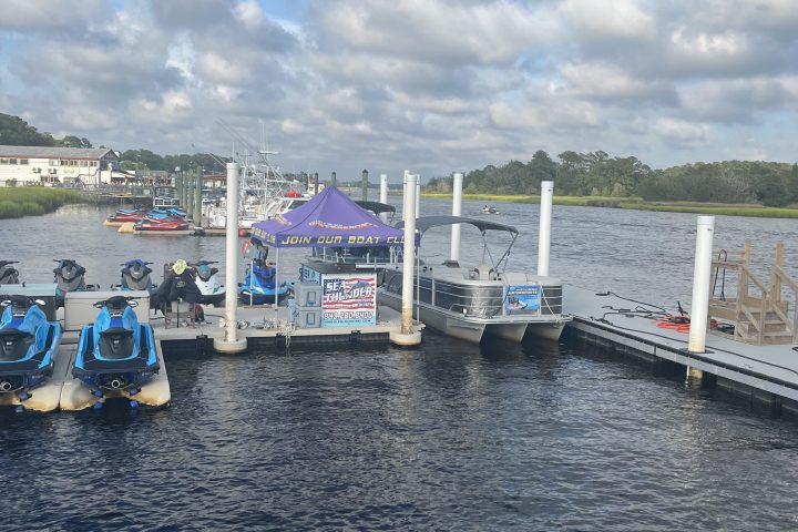 Dock with jet skis and boats under cloudy sky near a riverbank.