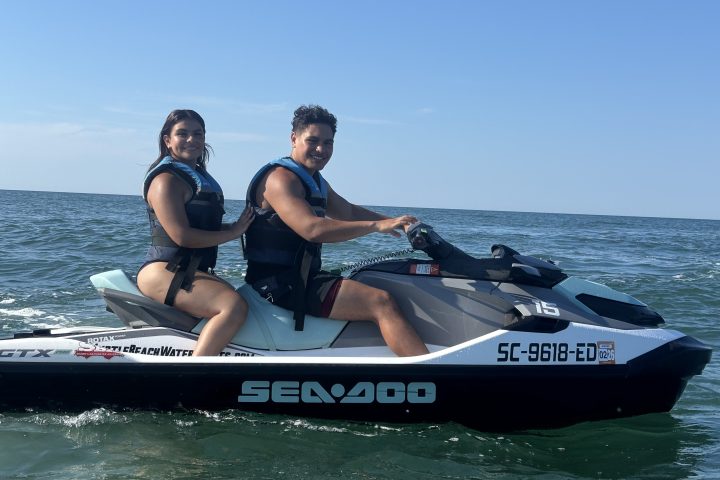 Two people riding a jet ski on the ocean, wearing life jackets under a clear blue sky.